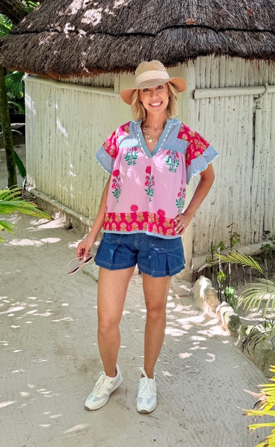Woman in a colorful blouse and denim shorts standing in front of a thatched-roof structure.