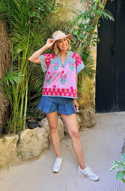 Woman in colorful embroidered top and denim shorts standing outdoors with plants and a stone wall in the background.