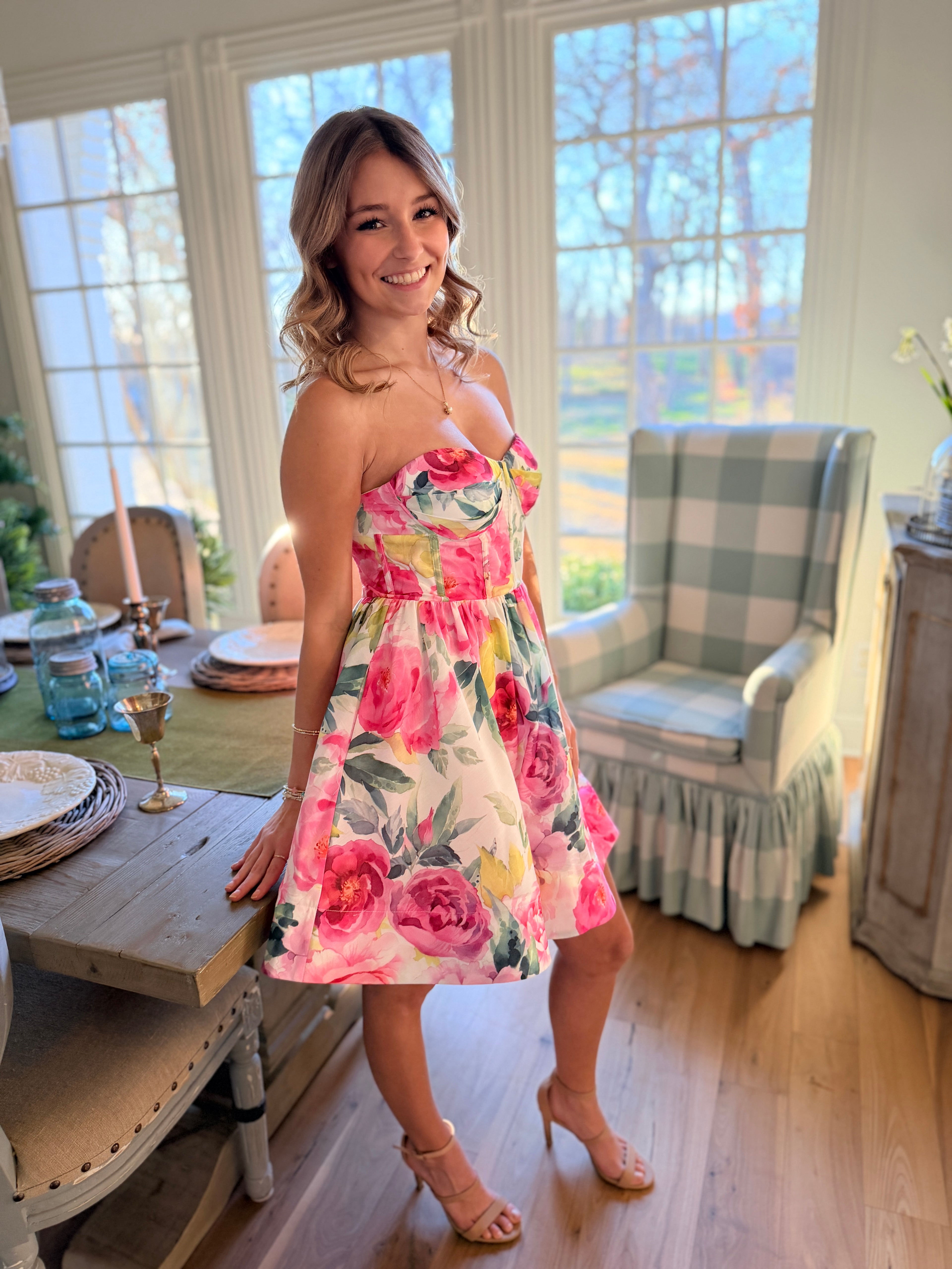 Woman in a floral dress standing in a bright room with large windows.