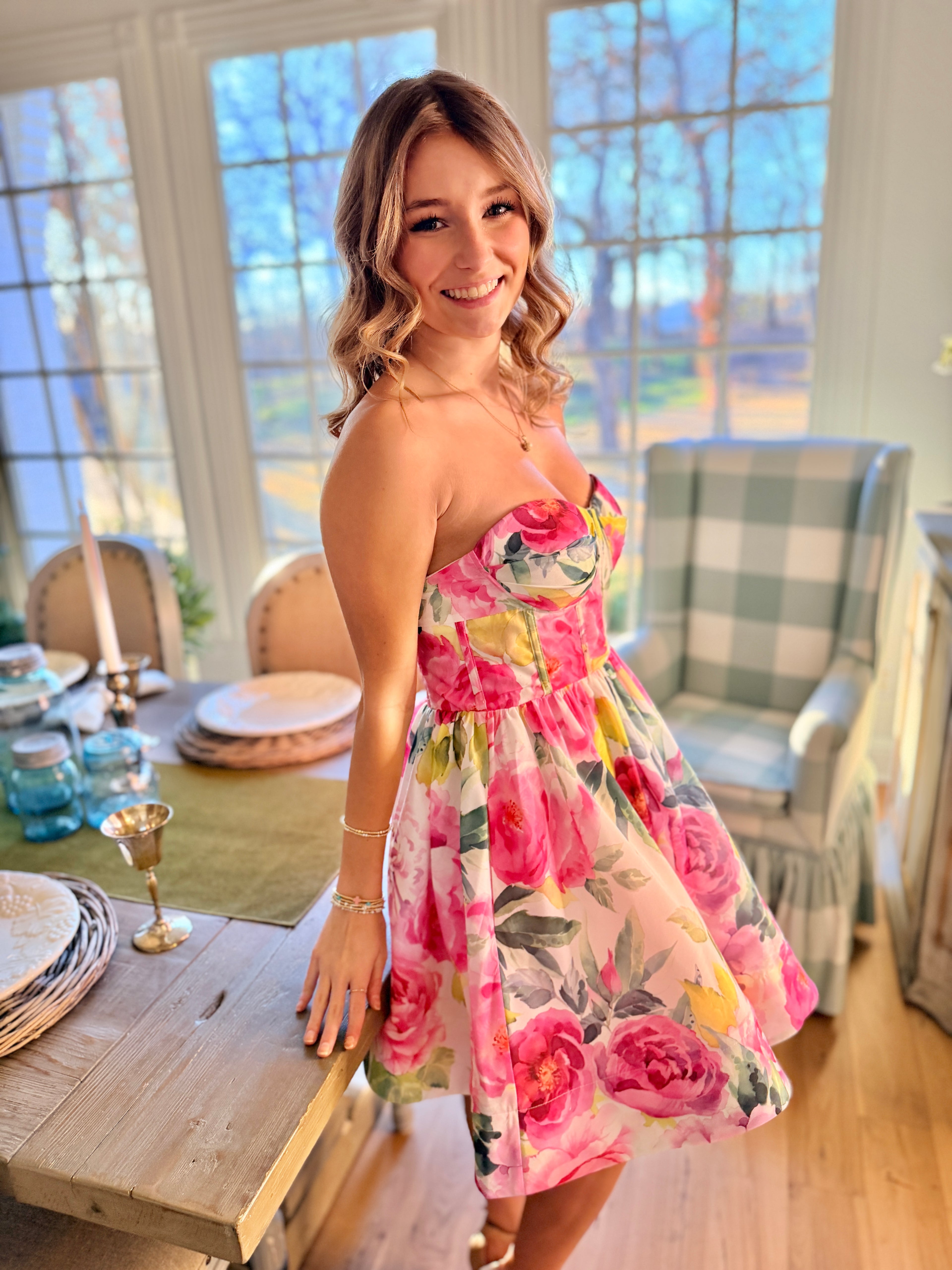 Woman in a floral dress standing in a bright room with large windows.