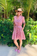 Woman in a pink patterned dress and hat standing in a tropical setting with palm trees.