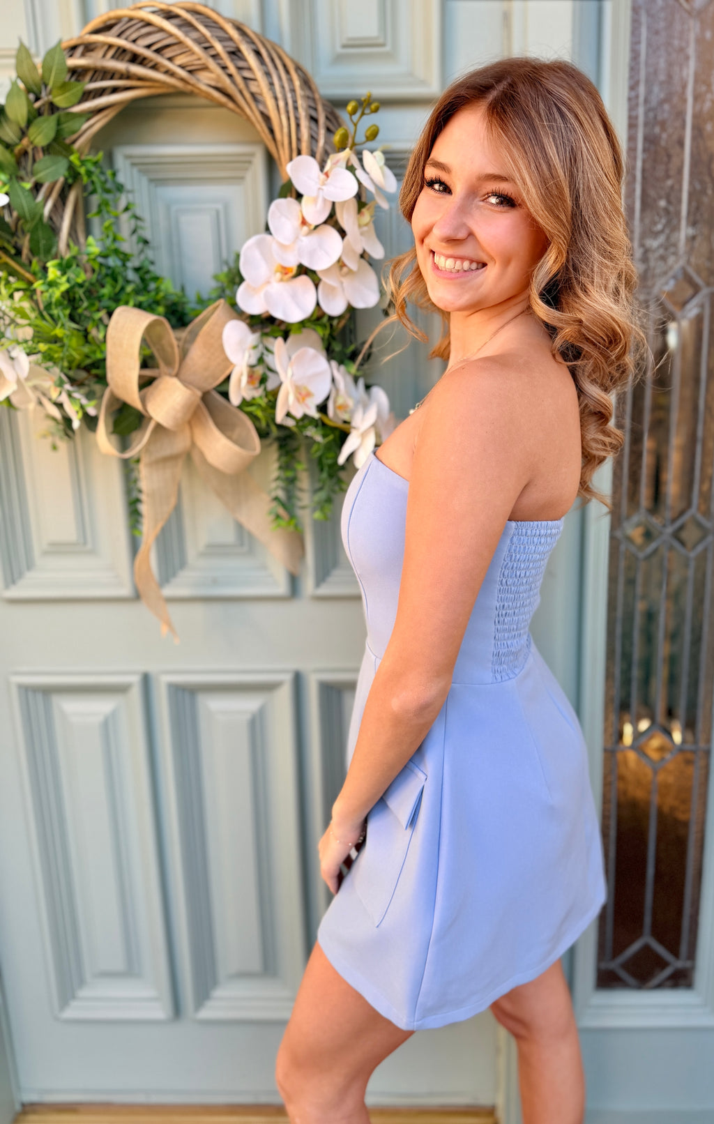 Woman in a light blue dress standing in front of a door with a decorative wreath.