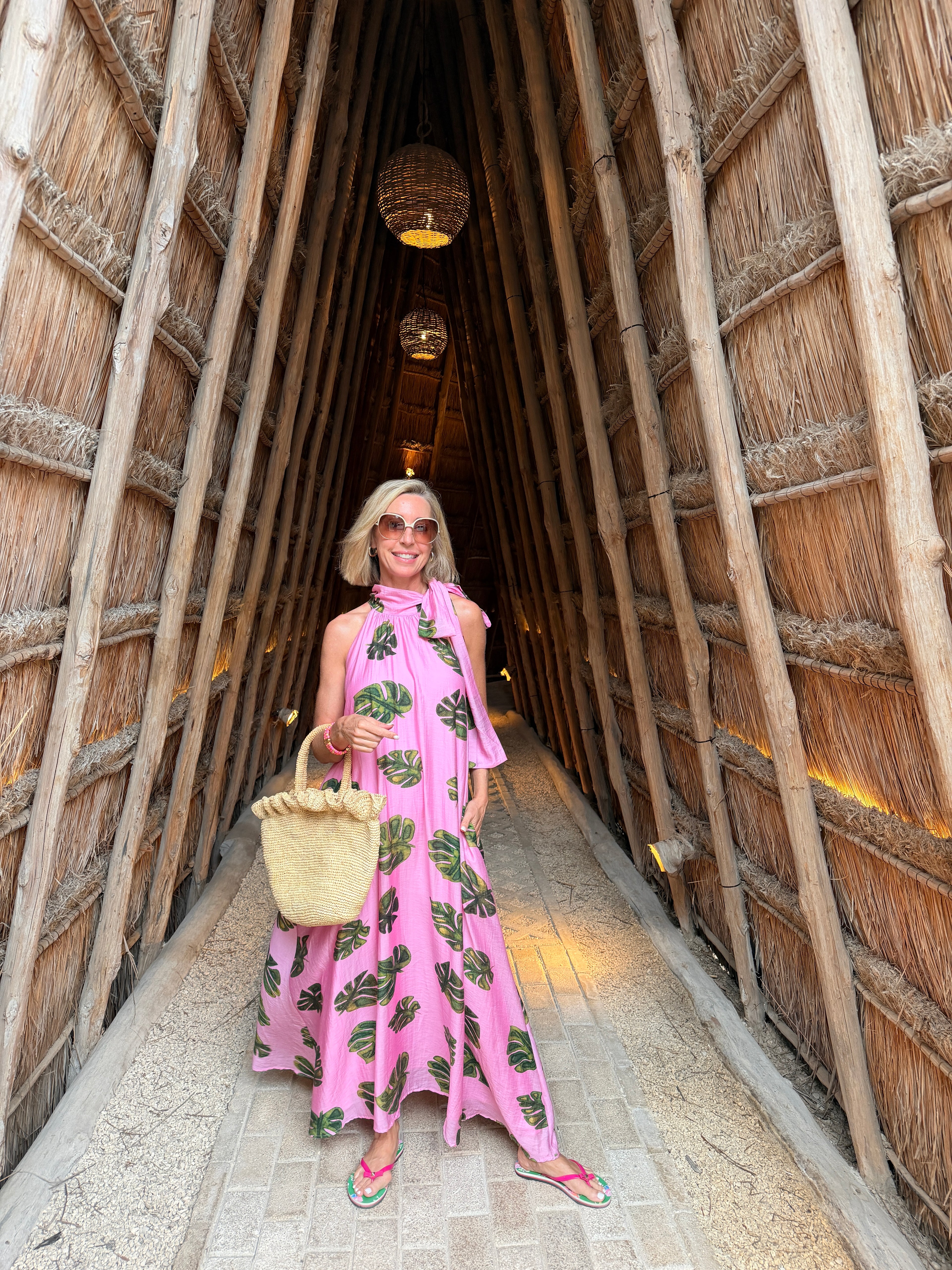 Person in a pink dress with green patterns standing inside a thatched-roof structure.